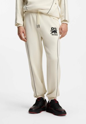 Man draagt crème joggers met zwarte bies, RB Hugo-logo op linker dij, gecombineerd met zwarte sneakers met rode accenten.
