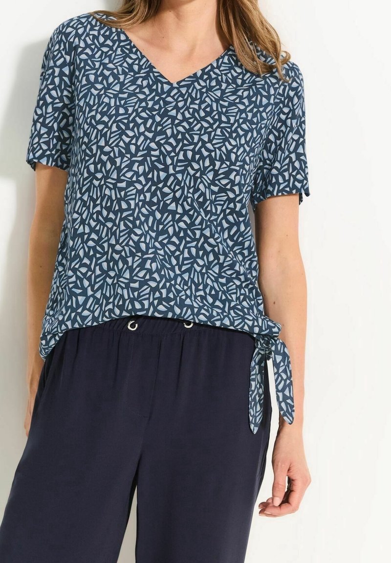 Femme portant un haut bleu à manches courtes à motifs avec col en V et lien sur le côté, ainsi qu'un pantalon ample bleu marine foncé sur fond blanc.
