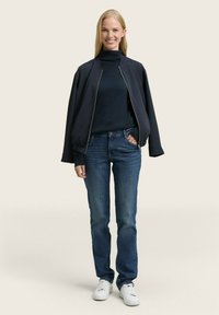 Veste de bombardier marine portée sur un turtleneck noir, avec un jean bleu et des baskets blanches. La veste possède une fermeture éclair à l'avant et des poignets élastiques.