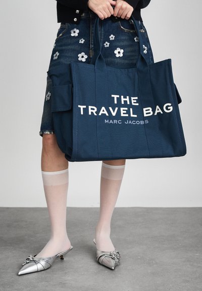 Bolsa tote azul escuro com grande texto branco "THE TRAVEL BAG MARC JACOBS", apresentando alças robustas e um bolso lateral. Usada com sapatos prateados e meias leves.