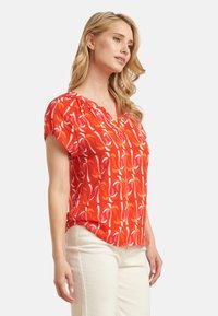 Rode blouse met witte en roze spiraalpatronen, korte mouwen en een V-hals ontwerp. De stof lijkt licht en soepel te zijn.