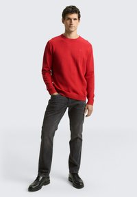 Maglione rosso lavorato a maglia con scollo classico rotondo, abbinato a jeans scuri e scarpe nere, esposto su uno sfondo grigio chiaro.
