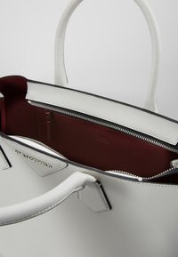 Sac à main en cuir blanc avec un design structuré, présentant un intérieur rouge profond, une poche zippée et des poignées doubles. Accent de logo argenté.