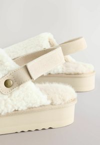 Sandales plateformes avec une tige en fourrure crème, texture douce et brides en daim beige. Présente un bouton-pression doré rond et une semelle à motif.