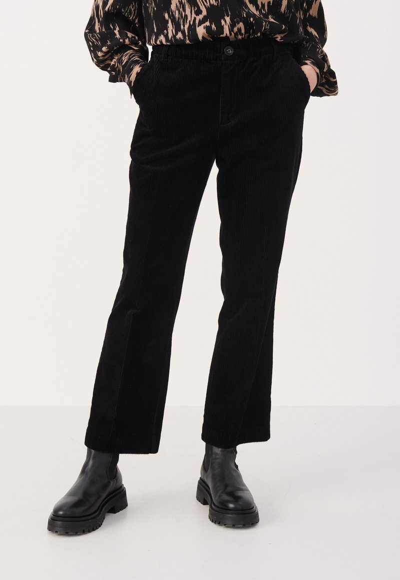 Personne portant un pantalon en velours côtelé noir, des bottes noires épaisses et un haut à manches longues imprimé animal, debout les mains dans les poches.