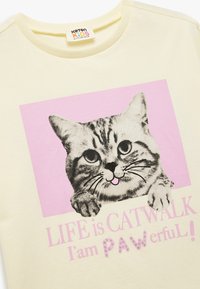 Vaalean keltainen puuvillapaita, jossa on grafiikka harmaasta tabbykissasta vaaleanpunaisella taustalla, ja teksti "LIFE on CATWALK, olen PAWerful!" vaaleanpunaisella.