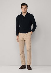 Man som bär en svart kvartdraget tröja, beige byxor och bruna kostymskor står med en hand i fickan mot en enfärgad bakgrund.