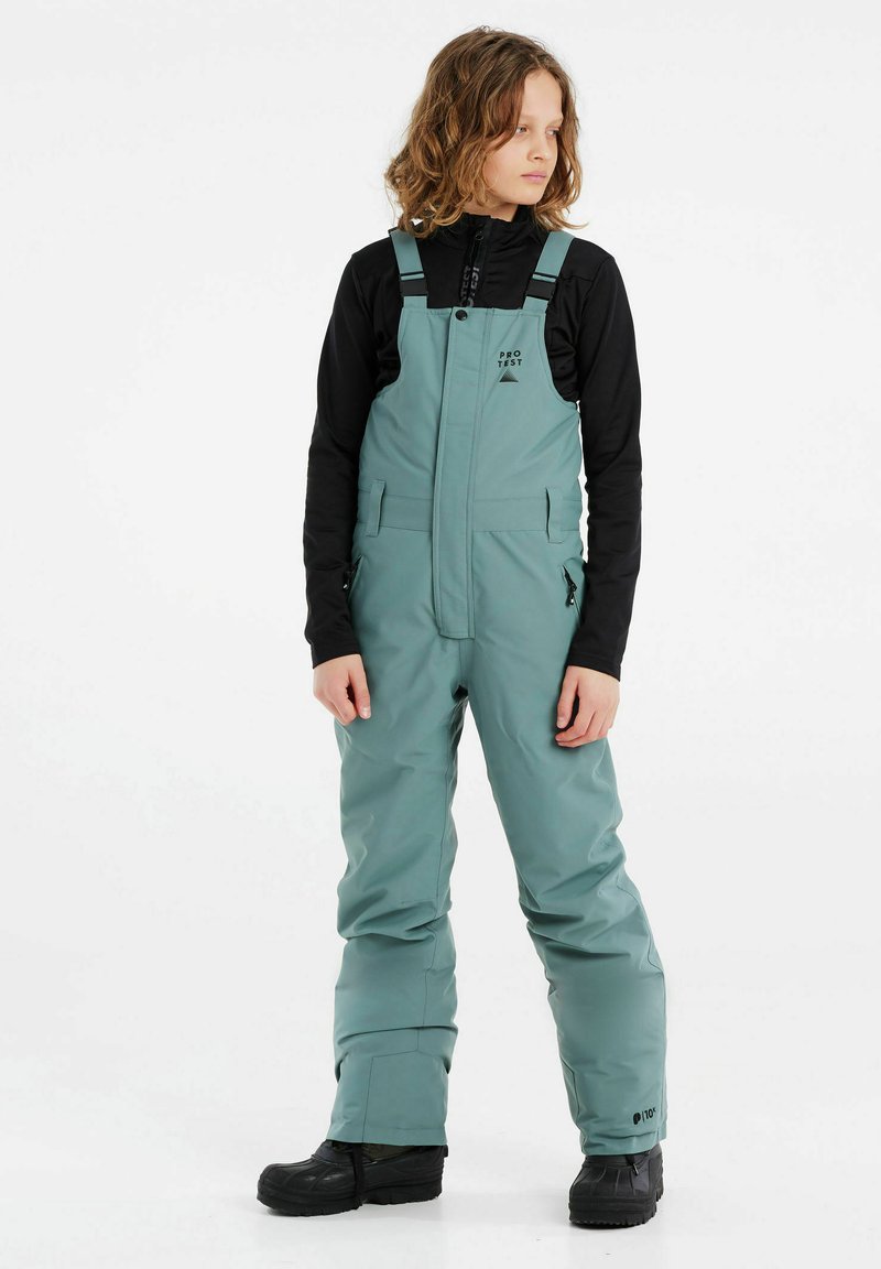 Salopettes vert eau avec bretelles réglables, poches zippées et coupe décontractée. Portées par-dessus un basique à manches longues noir et assorties à des bottes noires.