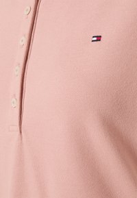 Jasnoróżowa koszulka polo z białymi guzikami i małym prostokątnym logo w kolorach czerwonym, białym i granatowym na klatce piersiowej.