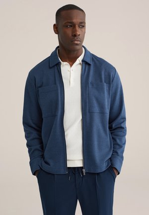 Marineblauwe jas met ritssluiting, twee borstzakken, gestructureerde stof en een losse pasvorm, gedragen over een wit overhemd met kraag.