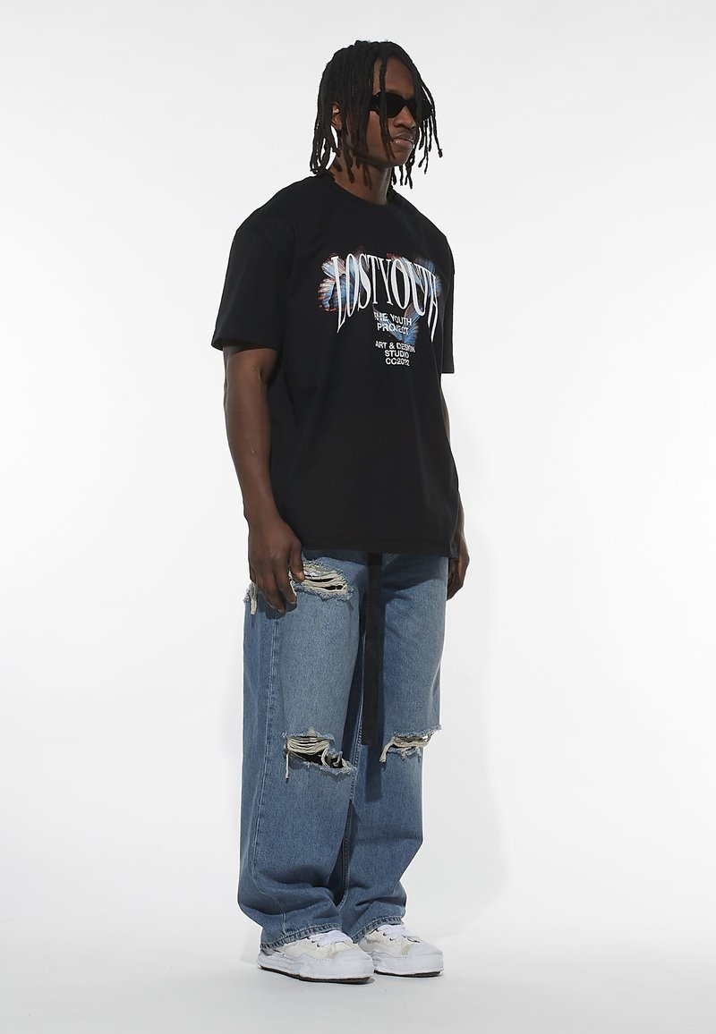 Sort grafisk t-shirt med tekstdesign, parret med slidte blå jeans og hvide sneakers. Model iført solbriller, står mod en hvid baggrund.