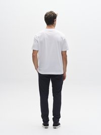 Mees, kes kannab valget lühikeste varrukatega T-särki ja tumesiniseid pükse, vaadates tagantpoolt, mustade tossudega ühtlase taustaga.