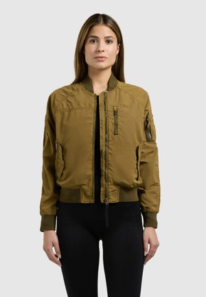 Femme aux cheveux bruns raides portant une veste bomber vert olive sur des vêtements noirs, debout devant un fond clair uni.