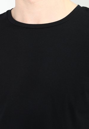 Persona vistiendo una camiseta negra de cuello redondo lisa, mostrando el escote y la parte superior del pecho sobre un fondo claro.