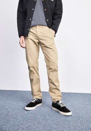Chinos beiges avec une coupe droite, associés à un t-shirt gris et une veste matelassée noire. Baskets noires avec des accents blancs.