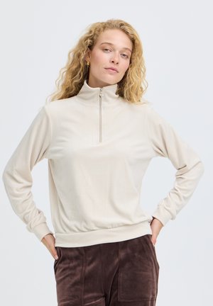Un modèle pose les mains sur les hanches, vêtu d’un sweat-shirt crème à fermeture éclair courte et d’un pantalon marron foncé, sur un fond clair uni.