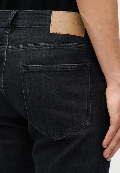 Το δεξί χέρι ακουμπά δίπλα σε μαύρο τζιν denim με δερμάτινη ετικέτα Tiger of Sweden πάνω από την πίσω τσέπη.