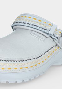 Sabot Crocs gris clair avec coutures décoratives jaunes et grises et une bride en tissu réglable sur le dessus.