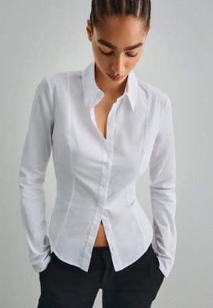 White cotton long-sleeve shirt with a fitted design, pointed collar, and button-down front. Smooth texture, slightly curved hem at the waist.