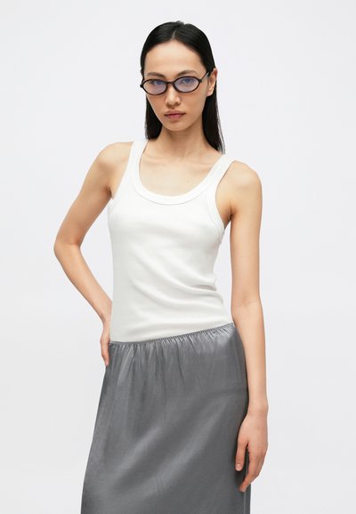 Maiou alb cu dungi, cu decolteu larg, asortat cu o fustă gri din satin, prevăzută cu o bandă elastică în talie. Modelul pozează cu o mână pe șold.