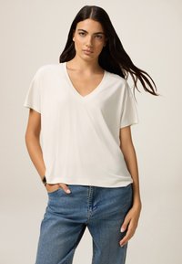 Hellbeige V-Ausschnitt-T-Shirt mit kurzen Ärmeln, weicher Textur, lockerer Schnitt, getragen mit blauen Denim-Jeans, minimalistisches Design.