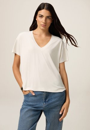 Hellbeige V-Ausschnitt-T-Shirt mit kurzen Ärmeln, weicher Textur, lockerer Schnitt, getragen mit blauen Denim-Jeans, minimalistisches Design.