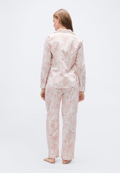 Γυναίκα που στέκεται ξυπόλυτη, φορώντας ανοιχτό ροζ πυτζάμα με floral σχέδιο, μπλούζα με μακρύ μανίκι και αντίστοιχο παντελόνι, γυρισμένη με την πλάτη προς απλό φόντο.