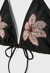 Crni bikini top s ukrasima od ružičastih i bijelih perli u obliku cvijeta i tankim podesivim trakama na donjem srednjem dijelu.