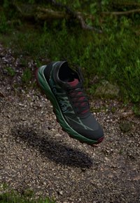 Schwarzer und grüner Sportschuh mit einer strukturierten Oberfläche, roten Akzenten und einer robusten Sohle, der über einem Schotterweg zu schweben scheint.
