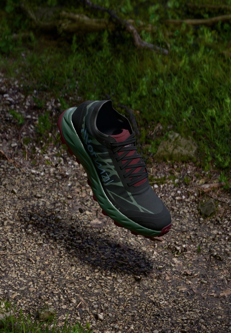 Schwarzer und grüner Sportschuh mit einer strukturierten Oberfläche, roten Akzenten und einer robusten Sohle, der über einem Schotterweg zu schweben scheint.