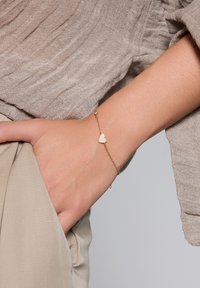 Złota bransoletka z serduszkiem ozdobionym przezroczystymi cyrkoniami, noszona na nadgarstku, z teksturowanym materiałem w tle.