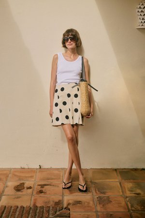Vrouw in wit mouwloos topje, beige rok met grote zwarte stippen, zwarte teenslippers, grote zonnebril en een geweven schoudertas.