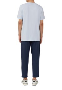 Persoon in een lichtblauw shirt met korte mouwen, donkerblauwe broek en witte sneakers, staand met de rug naar de camera tegen een witte achtergrond.