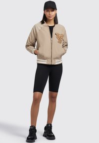 Beige bomberjack met een geborduurd patch, ritsluiting, witte gestreepte boorden, gedragen over een zwarte outfit en gecombineerd met zwarte sneakers.