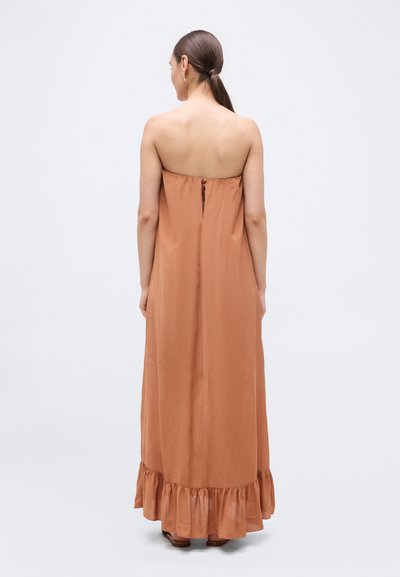 Kvinde med brunt hår i en lav hestehale iført en lang, terrakotta farvet stropløs kjole med knapper bagpå og flæsekanter nederst, stående mod en neutral baggrund.