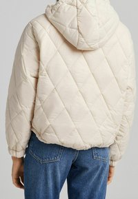 Béžová prošívaná bunda s kapucí, s diamantovým vzorem a elastickými manžetami. Nenosí se na modré džíny. Hladká textura.