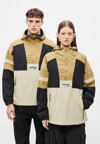 Δύο μοντέλα φορούν ελαφριά αντιανεμικά με κουκούλα σε μπεζ, μαύρο και καφέ χρώμα, με εμπρόσθιο φερμουάρ, ρυθμιστικά κορδόνια και διακριτικό λογότυπο στο στήθος.