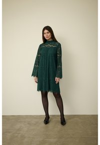 Zelené krajkové šaty s dlouhými rukávy, vysokým výstřihem a výstřihem ve tvaru oblouku. Nosené přes černé punčocháče a spárované s černými lodičkami se špičatou špičkou.