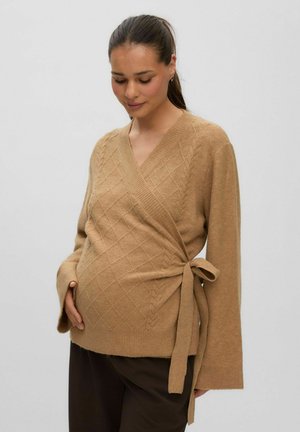 Femme enceinte tenant doucement son ventre, portant un pull cache-cœur marron clair avec un nœud sur le côté et un pantalon foncé.