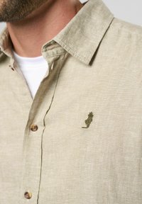 Beige linned skjorte med knap-ned krave, der har et grønt broderet detalje og brune knapper, der fremhæver dets teksturerede stof.