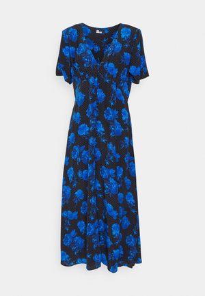 Femme portant une robe midi noire à imprimé floral bleu vif et manches courtes, associée à des bottines noires à talons.