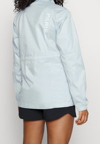 Ljusblå lättviktsjacka med en slät textur. Har vertikal text "PARIS" på ryggen och elastiska ärmslut. Bärs över svarta shorts.