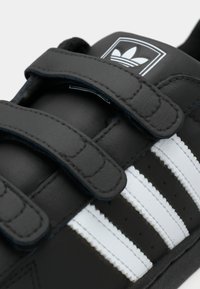 Sorte lær sneakers med to borrelåsremmer, hvite striper og en logo på tungen. Har en teksturert overdel med perforerte detaljer.