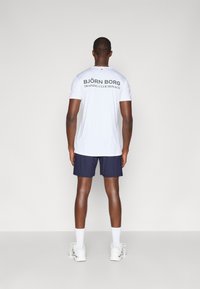 Λευκό κοντομάνικο πουκάμισο με την ένδειξη "BJÖRN BORG TRAINING CLUB MONACO" τυπωμένη σε σκούρο πράσινο στην πλάτη, συνδυασμένο με ναυτικές βερμούδες και λευκά αθλητικά παπούτσια.