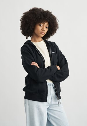 Młoda osoba o kręconych włosach, ubrana w czarną bluzę z kapturem na zamek błyskawiczny, na jasnej koszulce i jasnoniebieskich spodniach, stojąca z założonymi rękami.