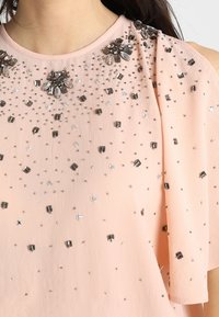 Světle růžová látka s rozptýlenými černými, stříbrnými a průhlednými korálkovými ozdobami, soustředěnými poblíž kulatého výstřihu a na ramenech.