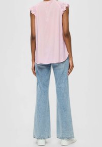 Personne portant un haut sans manches rose pâle avec des bords en dentelle festonnés et un jean large taille haute bleu clair, debout devant un fond uni.