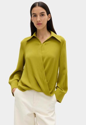 Vrouw met een mosterdgele blouse met lange mouwen en een brede kraag, gecombineerd met een witte broek, staand met handen in de zakken, neutrale achtergrond.