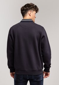 Granatowy sweter z okrągłym dekoltem, z teksturowanej tkaniny, z prążkowanymi mankietami i pasem. Posiada kontrastowy, paskowany detal na dekolcie.