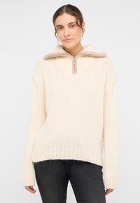 Kremowy sweter z dzianiny z wysokim kołnierzem, z częściowym zamkiem błyskawicznym i ściągaczem na hemie. Miękka tekstura, luźny krój i długie rękawy.